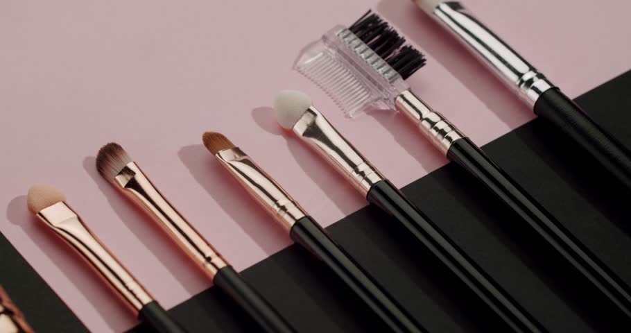 Professional makeup brushes set for applying foundation, eyeshadow, and powder lying on a pastel pink and black background in a slow motion dolly shot showing the beauty tools assortment