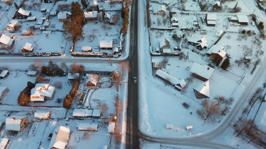 Aerial view shows a straight two lane road bisecting snow covered plots, compact houses, fenced yards, greenhouse rows, bare trees, and a solitary vehicle in warm morning light.