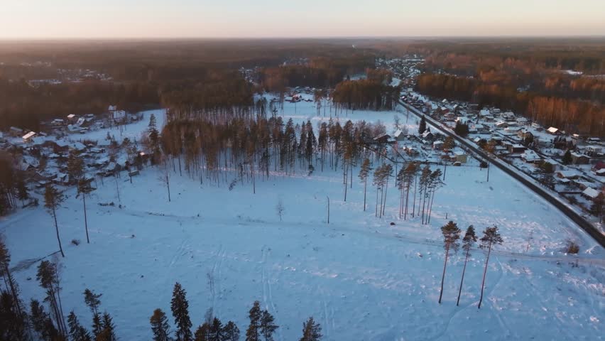 Aerial view shows snow fields, pine trunks, a straight road, and small houses. Warm low sun casts long orange highlights and shadows. Slow pan and drift reveal details.