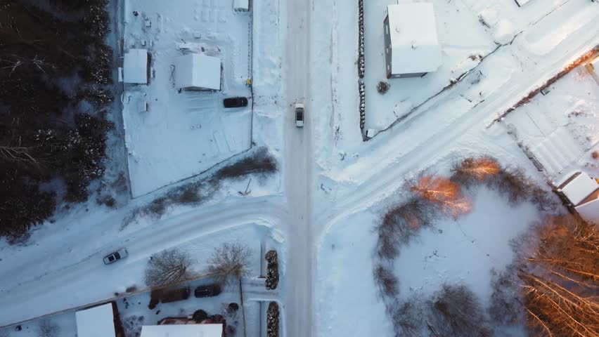 Aerial view of snow covered rooftops and a straight road bisecting a neighborhood. A single vehicle moves slowly as warm low sun casts long orange highlights.