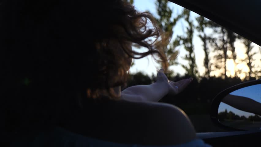 Back view of a young woman with curly hair enjoying the journey as she sticks her hand out of the moving car window, feeling the air and watching the sunset with a beautiful lens flare effect