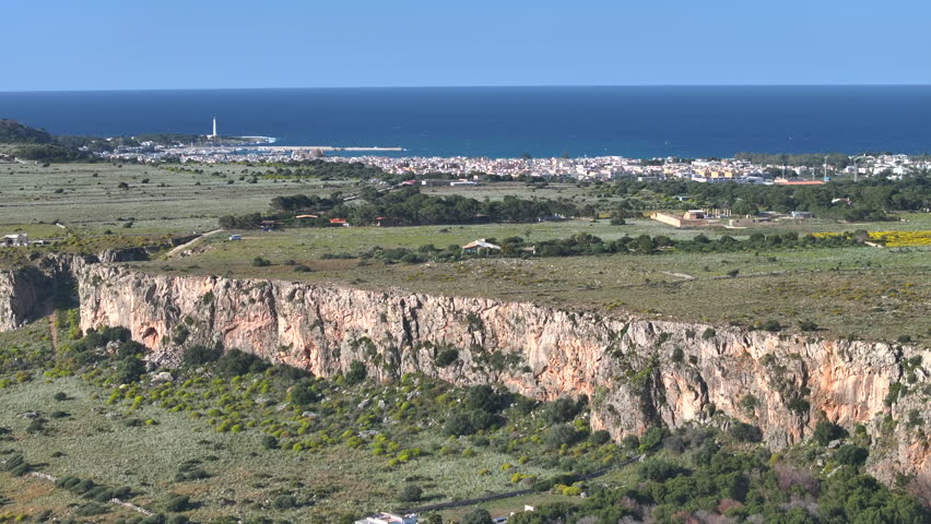 AERIAL: Long limestone cliff with a coastal town, a harbor and the blue Mediterranean Sea stretching out in the background. Picturesque coast of Sicily offers endless opportunities for rock climbing.