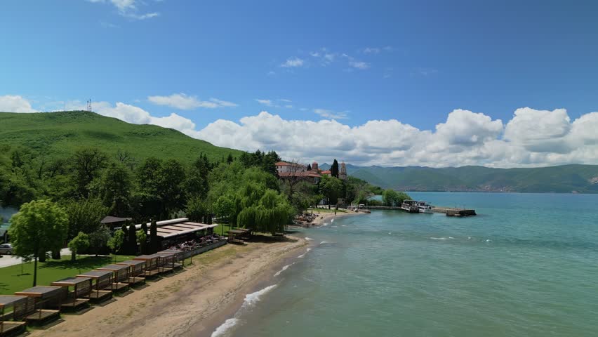 Aerial drone footage of Saint Naum Monastery on Lake Ohrid, North Macedonia. Scenic view of the historic Orthodox monastery, surrounded by clear blue water, lush greenery and rugged lakeside landscape. Iconic Balkan religious landmark and popular tourist destination.