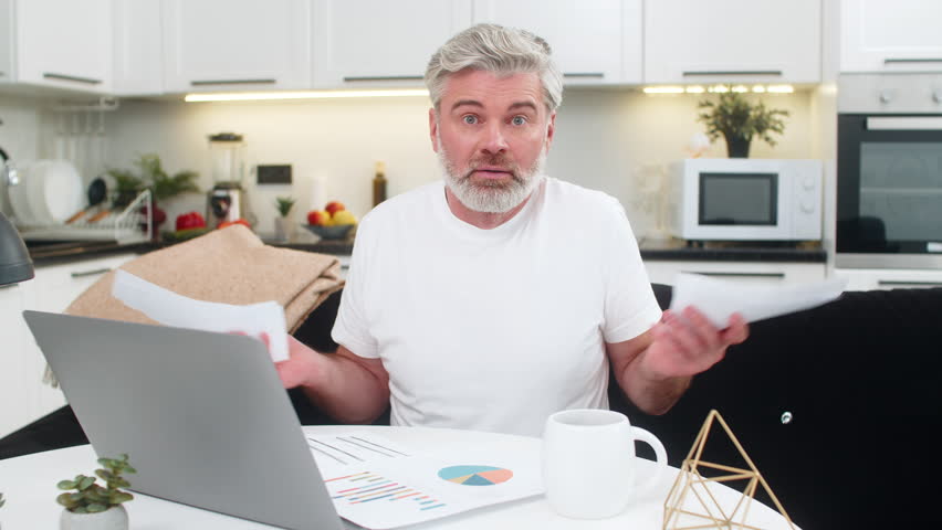 Middle-aged man at home reviews documents invoices and tax forms at table with laptop feels deeply lost stressed. Mature guy glances at camera confused by high sums wonders what broke needs support.