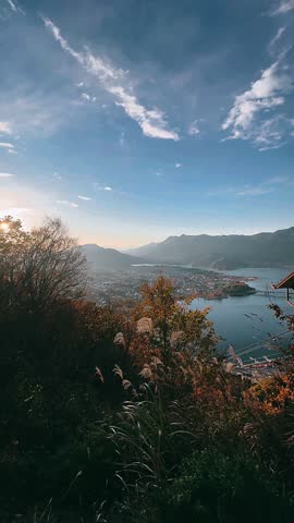 A wide hillside view overlooking a calm lakeside town framed by autumn grasses, colorful trees, distant mountains, and a soft blue sky with gentle clouds.
