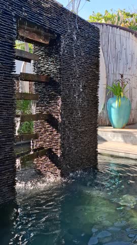 Water cascades down stacked dark stone slabs forming streams that fall into a pool below. Sunlight highlights the texture of the stones while greenery in the background creates a calm tropical garden atmosphere.