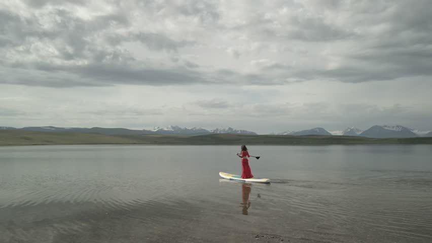 Raw drone footage follows at a close distance as a woman in a red dance dress stands on a paddleboard, gliding across a crystal-clear mountain lake with visible bottom, majestic snow peaks beyond.