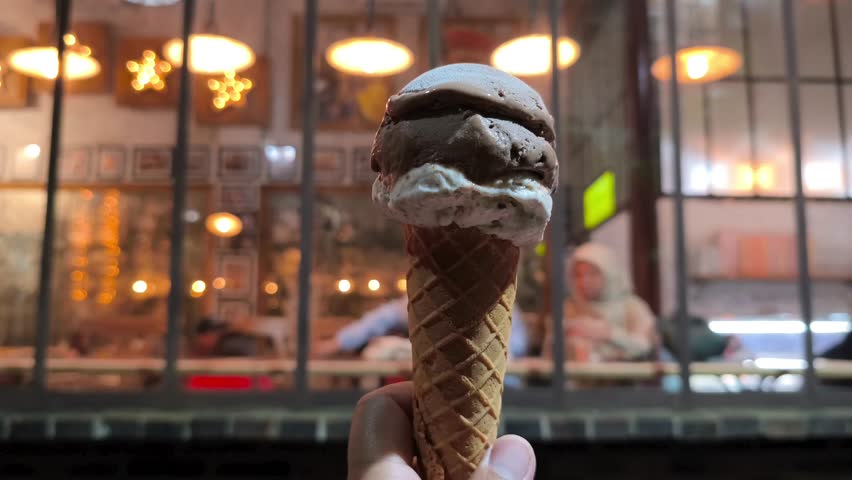 Hand holding chocolate and vanilla ice cream cone against cozy café lights at night background.