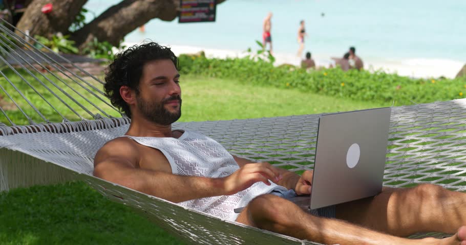Relaxed young Caucasian male tourist on vacation resting in beach hammock against the beach, talking with friends via video call on laptop. Online meeting and communication from anywhere in the world