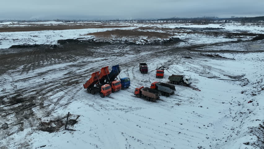Heavy trucks stand across snowy open field under gloomy sky. Industrial vehicles contrast with empty winter landscape and silence