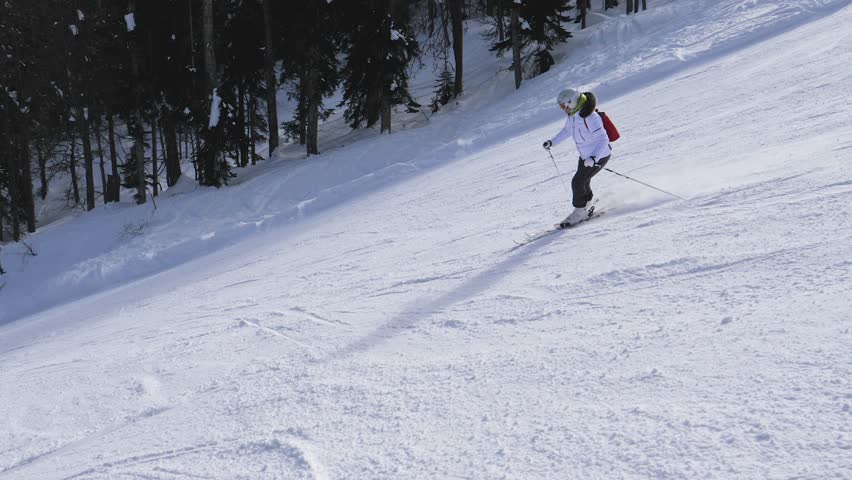 Professional female skier with backpack carving down groomed ski slope on winter day. Piste surrounded by dense pine forest. Skier enjoying active outdoor lifestyle and vacation