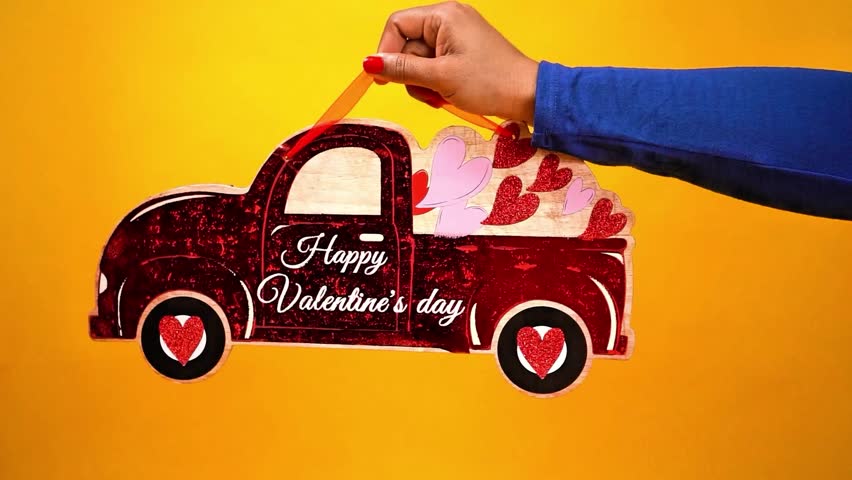 Close up of a hand holding a red Be My Valentine’s Day decorative truck filled with confetti hearts shape and a love message on a bright yellow background. Cheerful romantic concept with copy space, perfect for holiday promotions, greeting cards, social media, and love themed designs
