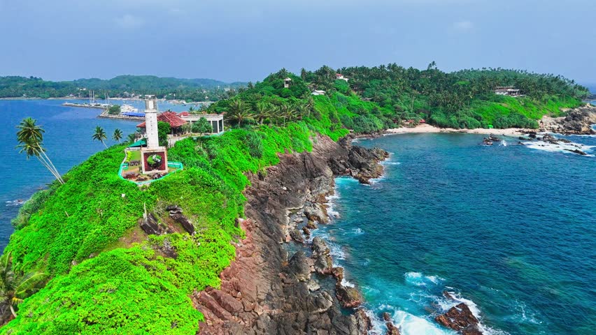 Sri Lanka - 05 05 2024: Drone view of a coastal lighthouse and small buildings on a lush green peninsula, surrounded by turquoise sea and palm trees under a bright sky.