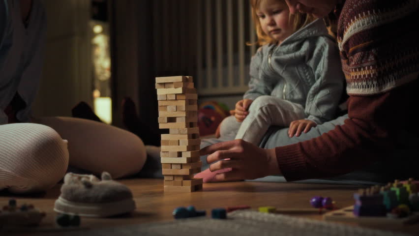 Mom, dad and daughter are playing board game sitting on the floor, they are stacking wooden blocks into high tower, enthusiastically playing in the warmth and comfort of their home, enjoying the time