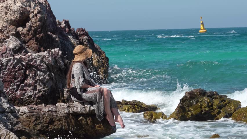 Woman sitting on rocks as ocean waves splash, slow motion