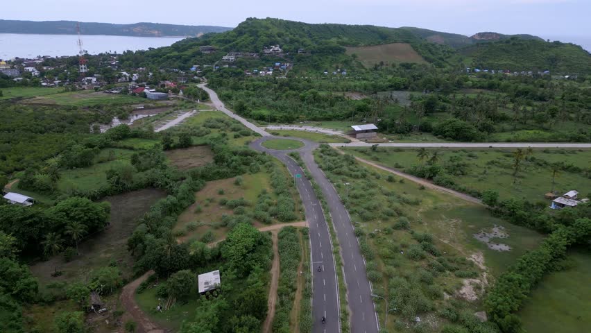 Aerial Coastal Crossroads Winding Road Village. Lush Hillside, Roundabout, Distant Coastline, Scattered Houses And Empty Fields. Scenes Include Farmer Tending Crops,Gerupuk,Lombok,Indonesia