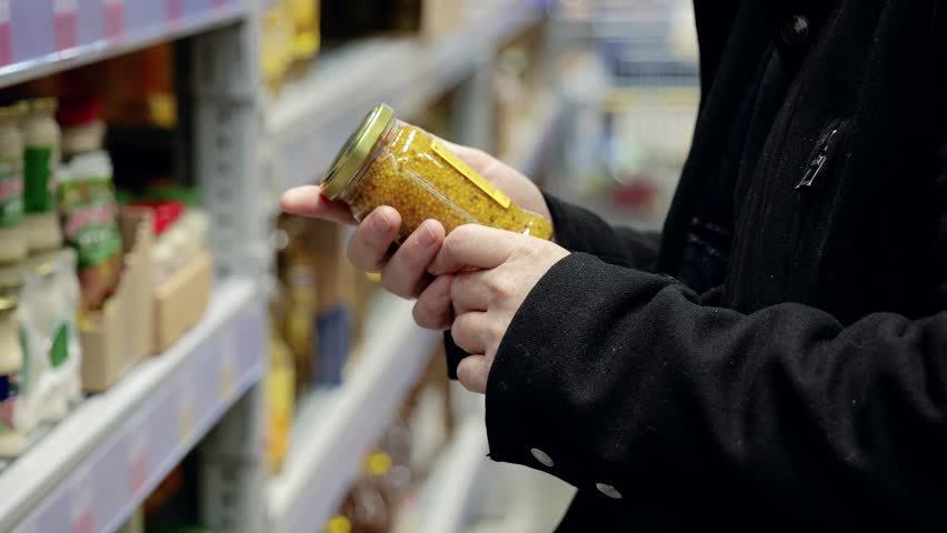Shopper checking french mustard jar, reading label closely while comparing product details in supermarket aisle during careful selection process