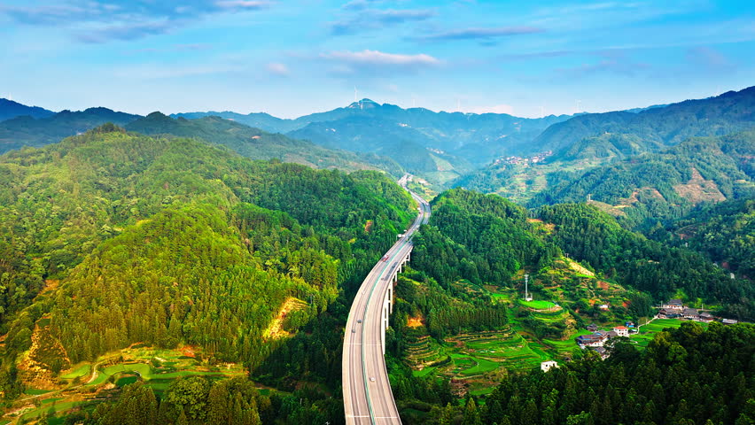 Aerial view of a modern highway bridge stretching through the dense green forests and mountain valleys in rural China. 