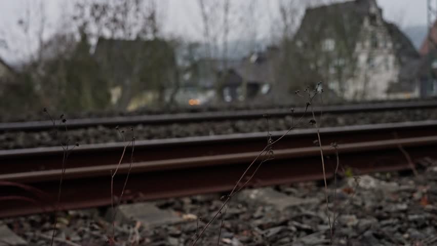 Curved Railway Tracks Through Industrial Edge, Grass And Gravel Fringe With Yellow Signal Box On Ballast, Overhead Catenary Masts And Bare Trees, Distant Depot Buildings Under Damp Winter Light, Mood