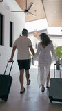 Rear view of a man and woman holding hands while walking with their luggage. The couple is arriving at a sunny tropical hotel for vacation. Vertical - Powered by Shutterstock - Get 15% off with code: PIKWIZARD15