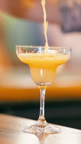 Close up of a bartender pouring a vibrant orange cocktail into a salt rimmed margarita glass, then a hand picking it up from the bar. Vertical