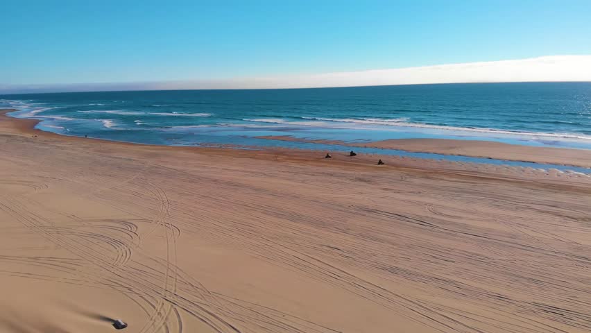 An aerial, wide-angle view of the sandy beach at Sand Lake, Oregon, with the blue Pacific Ocean waves rolling onto the shore under a clear blue sky. The scene captures the natural beauty and expansive
