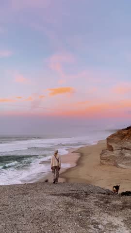 Woman with Dog Watching Ocean from Cliff at Sunset.

Woman and dog standing on cliff overlooking ocean, beach and pink sunset sky.