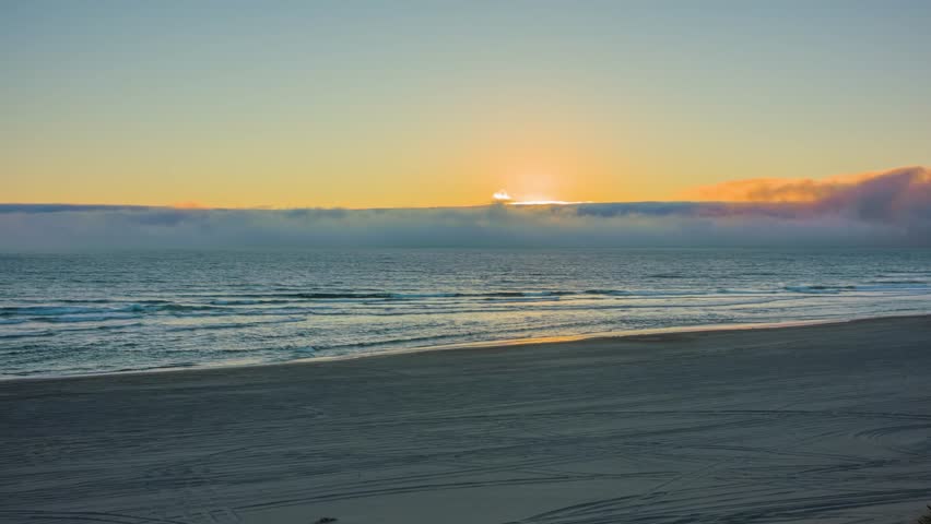 A tranquil and serene beach scene captures a golden sunrise or sunset over the ocean horizon, with the sun reflecting on gentle waves and wet sand. The sky transitions from a warm yellow-orange near t