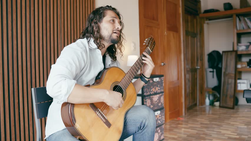 Talented musician with long curly hair passionately singing and playing a classic acoustic guitar. Male artist enjoying music in his room