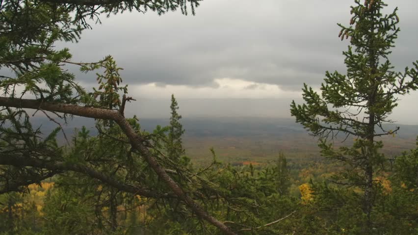 A scenic view overlooking an expansive autumn forest in the Southern Urals, Russia. The landscape features vibrant fall colors of yellow and green foliage under a dramatic, overcast sky.