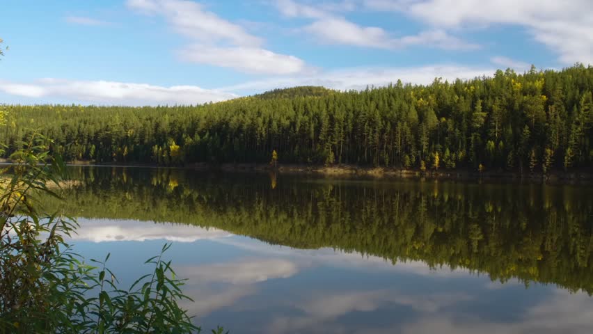 A tranquil natural landscape featuring dense green and yellow autumn forest trees reflected perfectly in the calm water of a lake under a blue sky with white clouds.