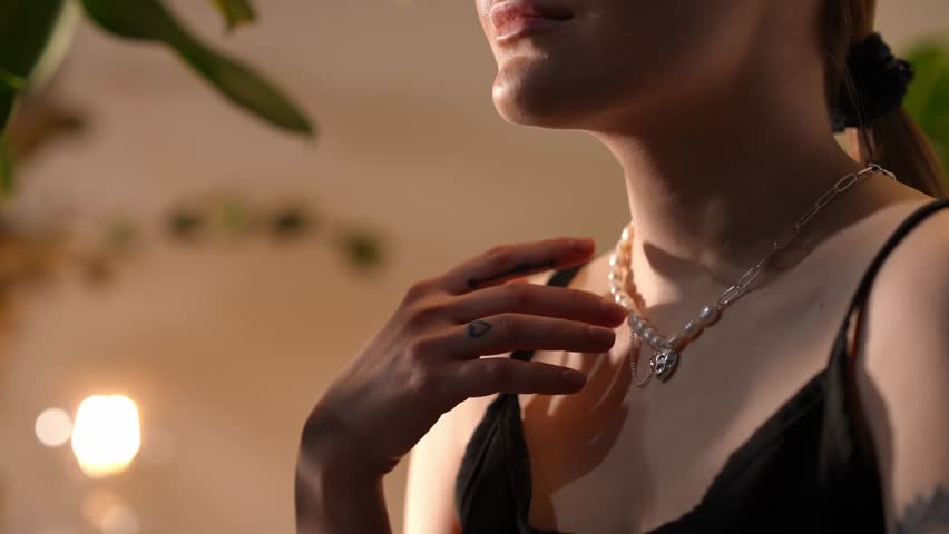 Young woman gently touches an elegant pearl and silver chain necklace on her neck. Close-up on the chest, neck, and hands with warm light