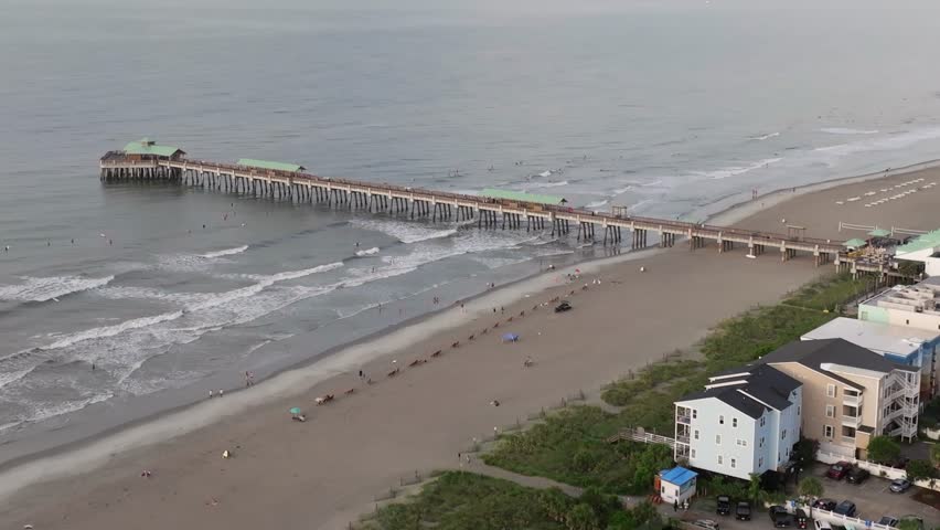 Aerial view of Folly Beach near Charleston, South Carolina lowcountry landscape showing beach and pier during morning sunrise with people enjoying the shore and surfers in ocean water - 4K Drone
