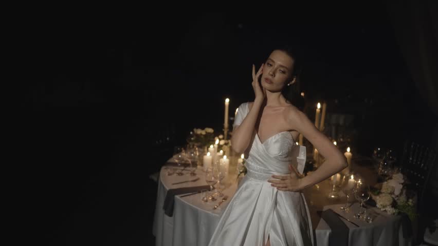 Beautiful bride in a white gown sensually posing in a dark room. Romantic wedding dinner setting illuminated by many warm candles