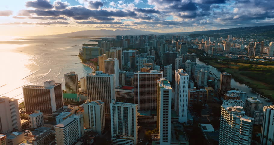Flight above Waikiki high-rises at sunset. Bright sun reflects from the surface of the ocean. Dark clouds over the city scenery.
