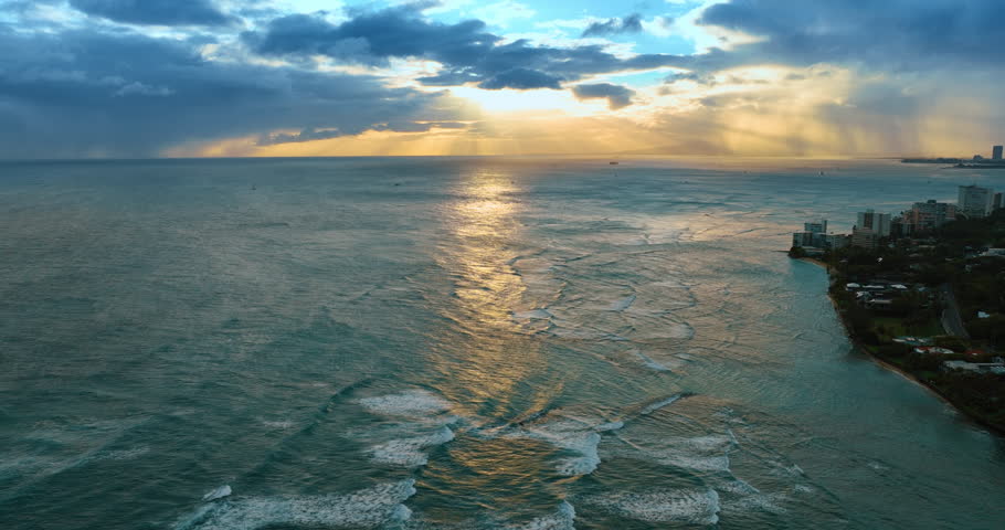 Rugged waterscape of the Pacific Ocean with waves rolling to the shore. Aerial view. Light of setting sun shines through the dark clouds. Hawaii, USA.
