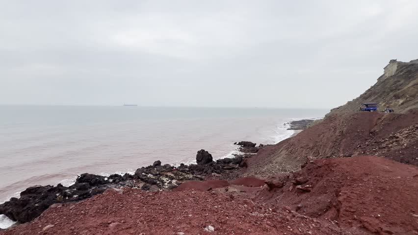 Red Soil Coastline and Rocky Shore with Pink Sea Under Overcast Sky
Wide shot of red soil cliff coastline and dark volcanic rocks by a calm pinkish sea under a cloudy sky. Natural coastal landscape background.