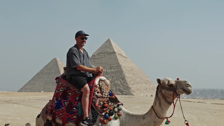 Classic Egyptian travel experience: a man sitting atop a camel at the Giza Plateau. The camel is adorned with a vibrant, traditionally patterned saddle.