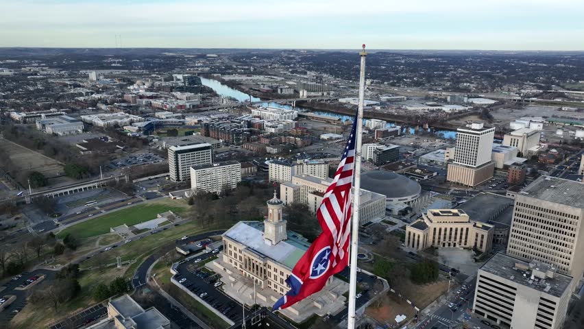 Top-down aerial view of Tennessee State Capitol in Nashville with American flag and Tennessee flag flying above city skyline during daylight.