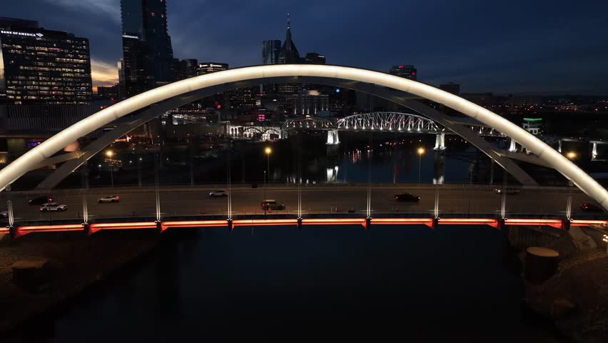 Aerial flight over modern illuminated bridge with driving traffic crossing river in Nashville, showing iconic downtown skyline at dusk transitioning into night. Passing by wide shot. Establishing shot