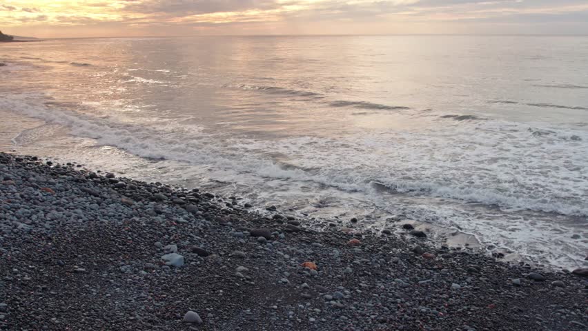 Calm ocean waves washing over a dark stone beach at dawn. Serene tropical coastline with soft sunlight, distant mountains, and cinematic water foam.