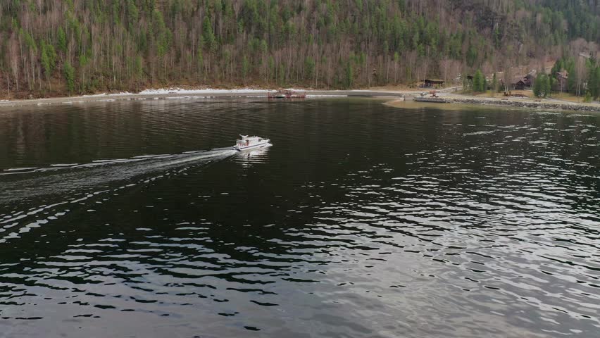 Shooting from a drone flying over a mountain lake, along which a speedboat is moving. A motorboat is rushing towards the hotel on the shore.