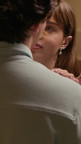 Vertical shot of tender young couple looking into each others eyes while slow dancing at home under warm light