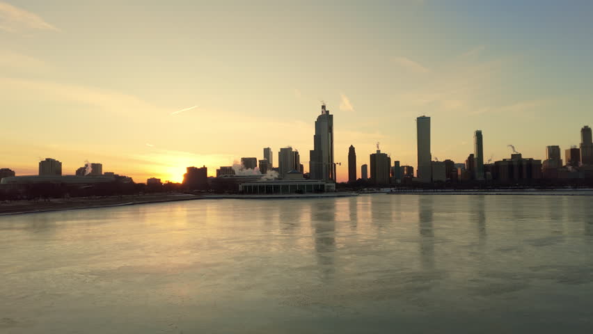 Chicago skyline with Lake Michigan at winter. Thin ice sheets and floating frozen formations cover water surface while warm golden light reflects off the city skyscrapers