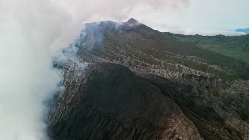 Drone aerial footage of Mount Bromo active volcano shows crater steam rising above rugged volcanic slopes, expansive caldera terrain and dramatic mountain landscape in East Java Indonesia.