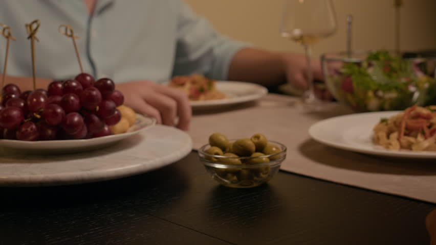 Handheld shot of young man and woman holding hands together, drinking white wine while dining at festive table during home celebration