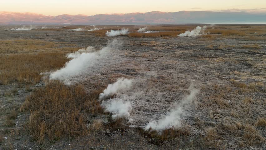 Low angle aerial shot of thick white vapor escaping from a geothermal vent in the middle of a brown field with distant mountains on the horizon during a cold morning