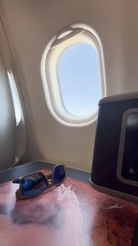 A pair of blue sunglasses sits on a wood-grain table next to an airplane window