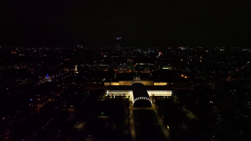 Aerial night view of a grand, symmetrically lit building standing out against the dark Paris cityscape. A modern skyscraper is visible on the distant horizon under a dark sky.