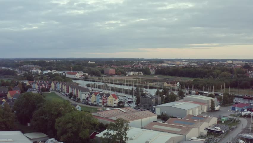 Aerial view of the Greifswald museum harbor on the river Ryck. Colorful gabled houses and numerous sailing ships along the quay define the maritime cityscape under a vast, cloudy sky.
